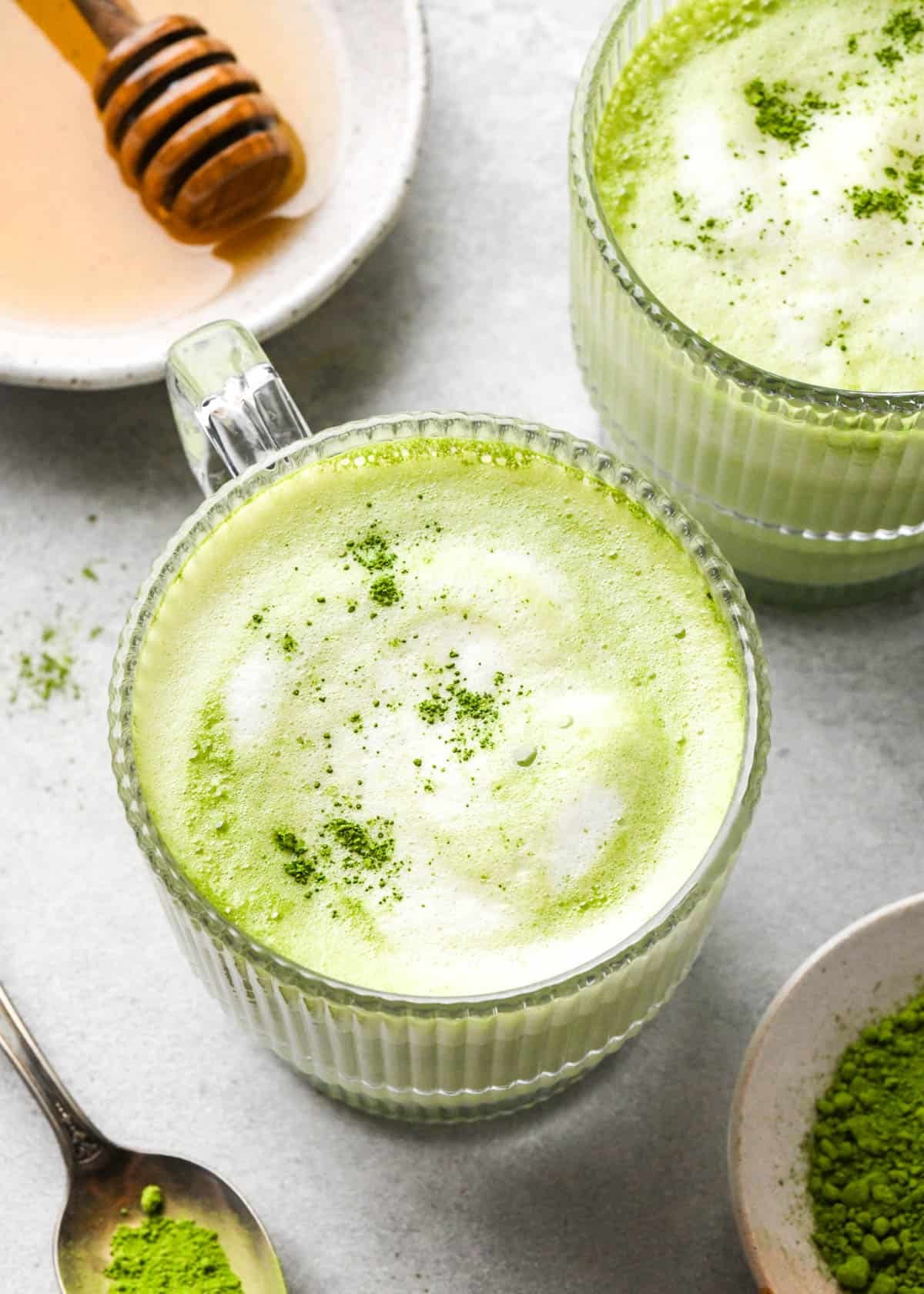 matcha latte in a clear glass mug with a handle with foamy milk on top sprinkled with matcha powder, and another matcha latte to the back and right of it