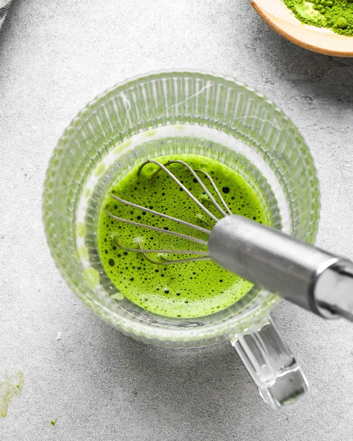 photo showing how to make a matcha latte - overhead view of hot water and matcha powder being whisked together in a glass mug
