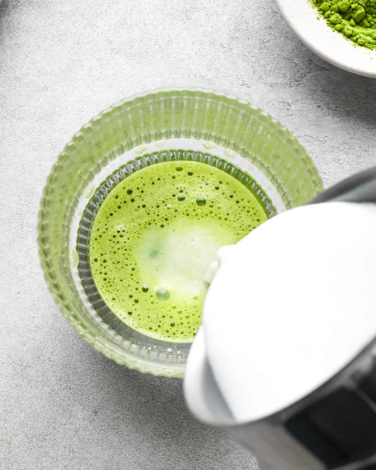photo showing how to make a matcha latte - overhead view of frothed milk being poured into a glass mug with matcha powder and hot water