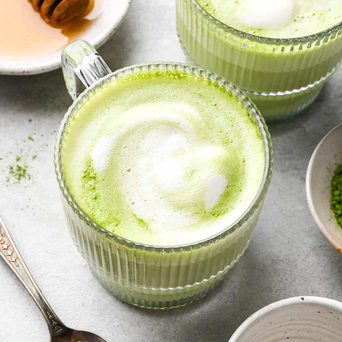 matcha latte in a clear glass mug with a handle with foamy milk on top and another matcha latte to the back and right of it