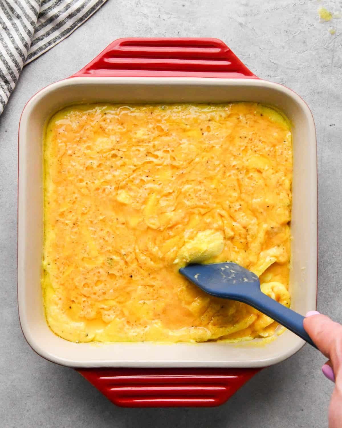photo showing how to make baked scrambled eggs - a blue spatula stirring the partially baked egg mixture in a red, square, ceramic baking dish
