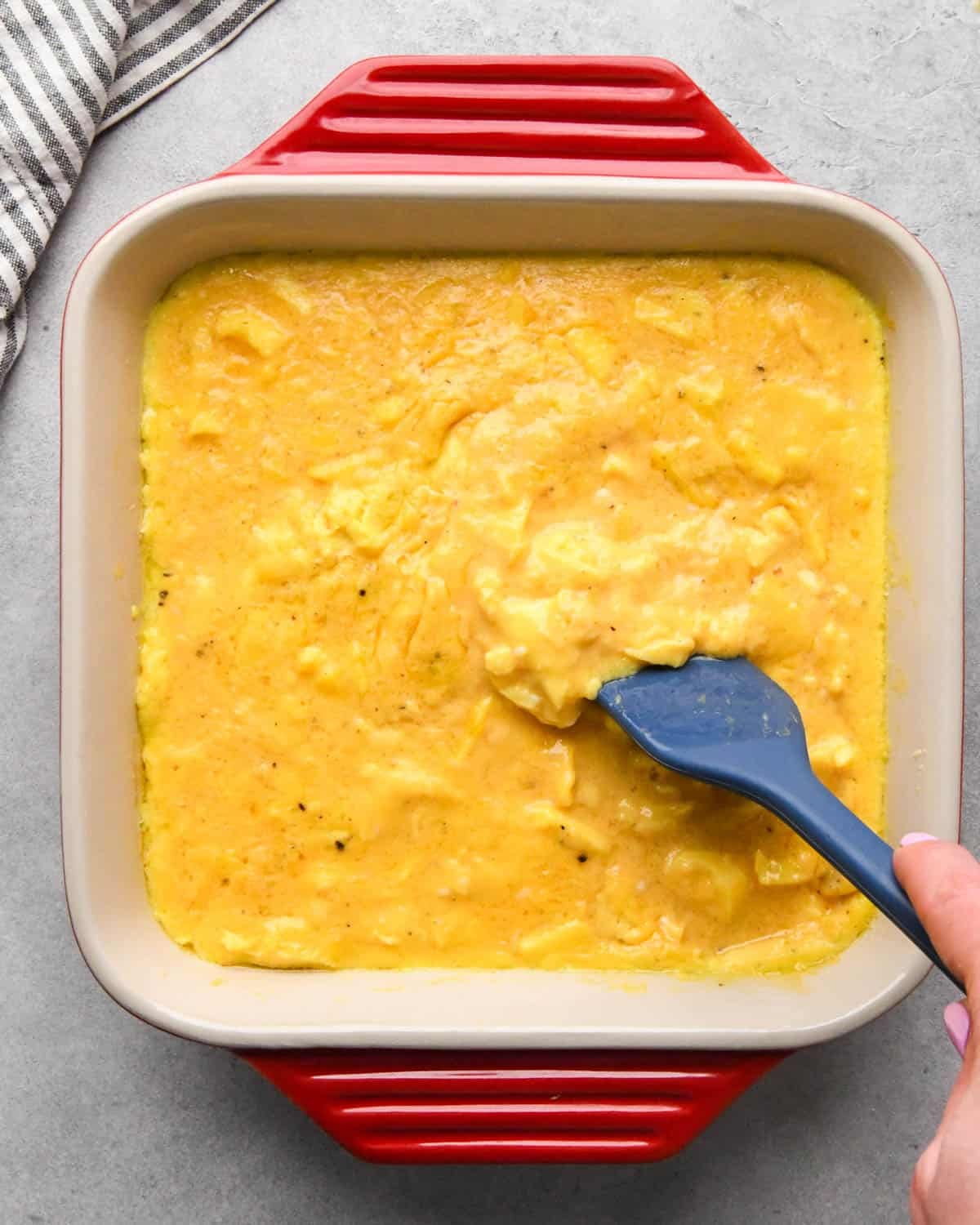 photo showing how to make baked scrambled eggs - a blue spatula stirring the partially baked egg mixture in a red, square, ceramic baking dish