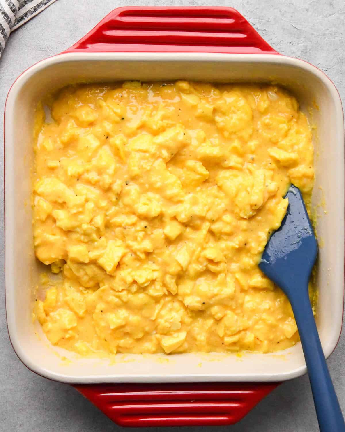 photo showing how to make baked scrambled eggs - partially baked egg mixture in a red, square, ceramic baking dish with a blue spatula after stirring before returning to the oven