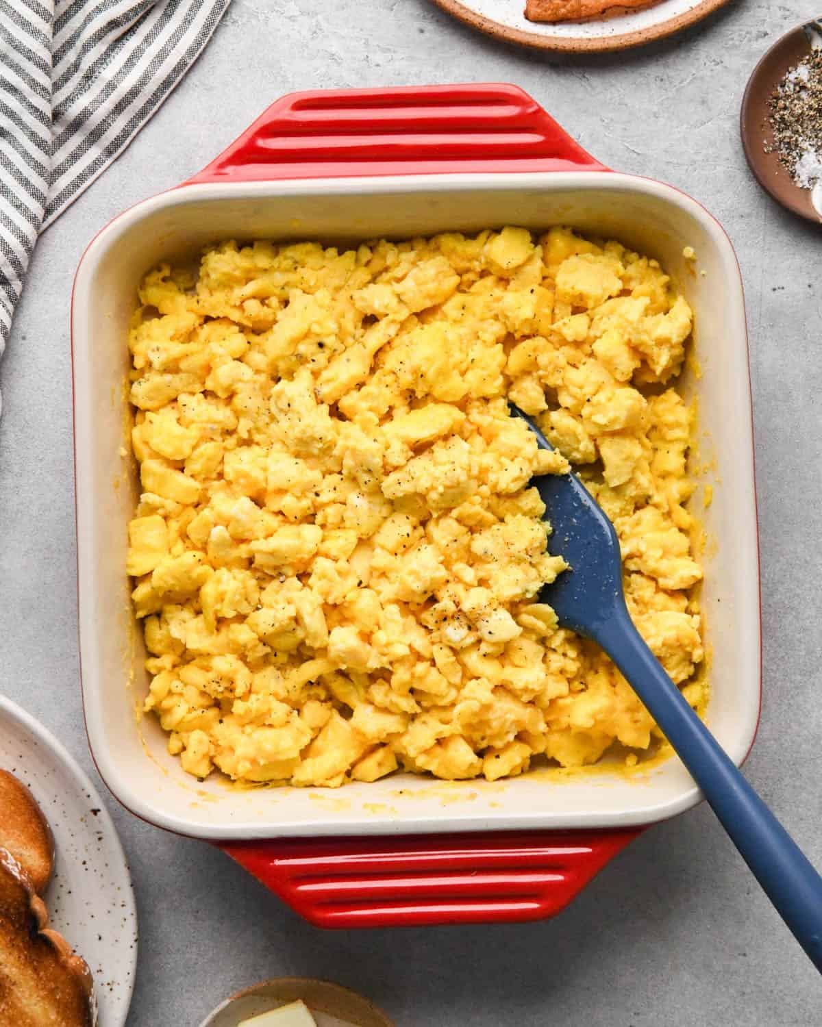 oven baked scrambled eggs in a red, square ceramic baking dish with a blue spatula in the eggs surrounded by toast and bacon