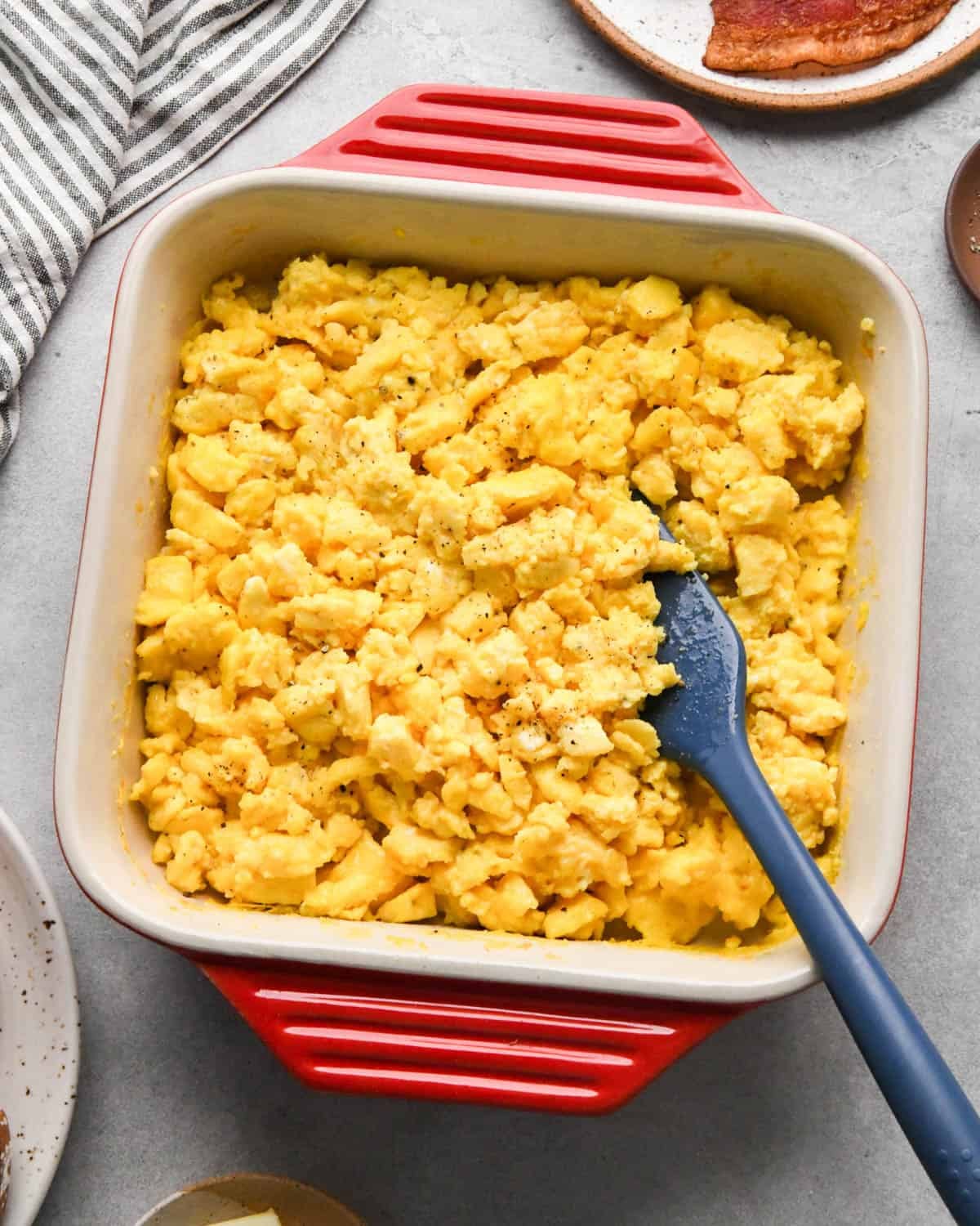 oven baked scrambled eggs in a red, square ceramic baking dish with a blue spatula in the eggs with a plate of bacon nearby