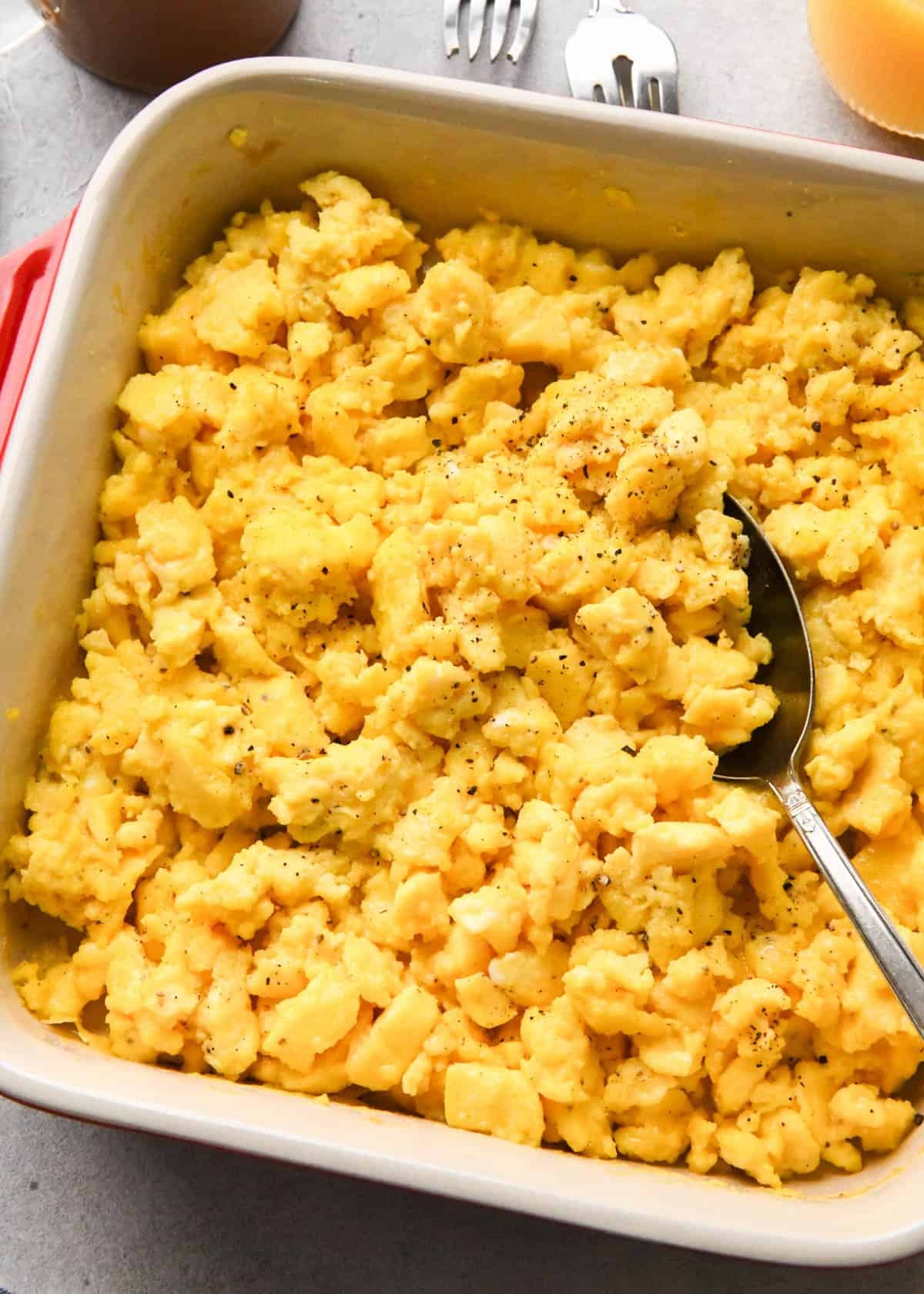 oven baked scrambled eggs in a red, square ceramic baking dish with a silver spoon in the eggs surrounded by coffee and orange juice