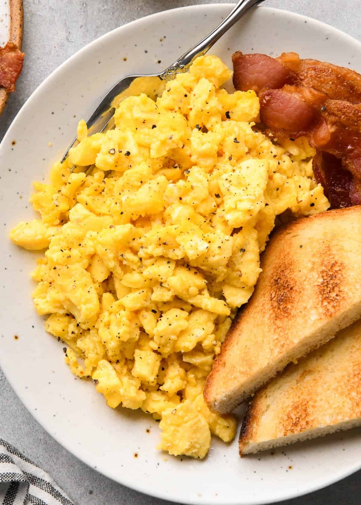 oven baked scrambled eggs topped with black pepper on a white plate with toast bacon and a fork.