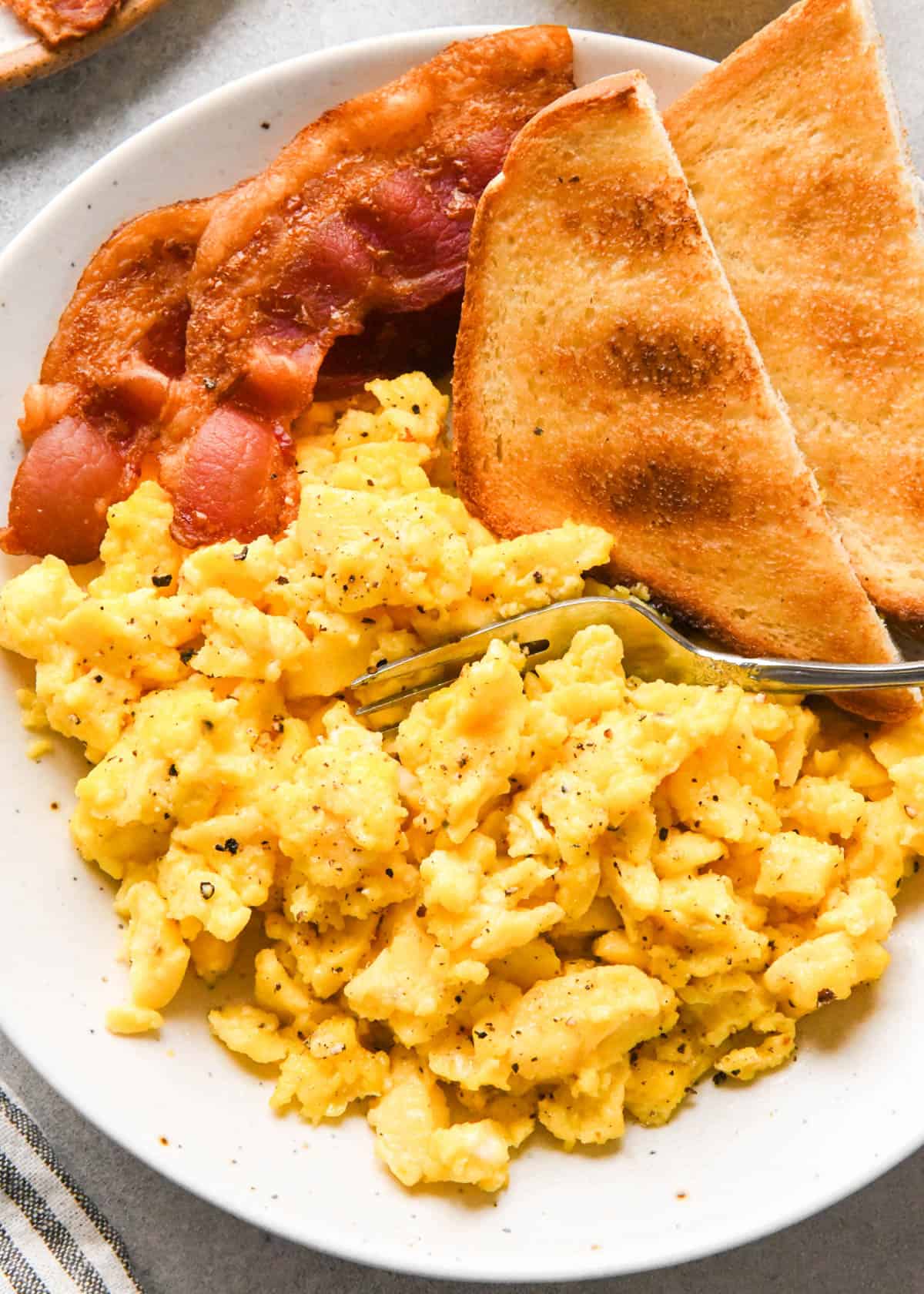oven baked scrambled eggs on a white plate with toast and bacon, there's a fork taking a bite of the eggs which are topped with pepper