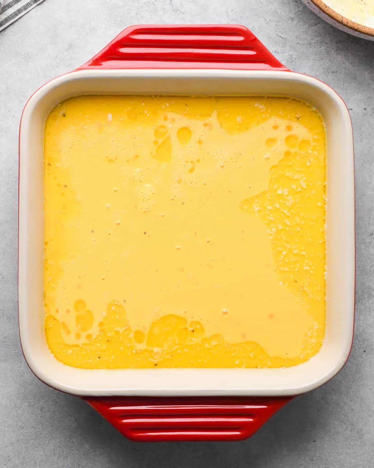 photo showing how to make baked scrambled eggs - whisked egg mixture poured into a red ceramic square baking dish
