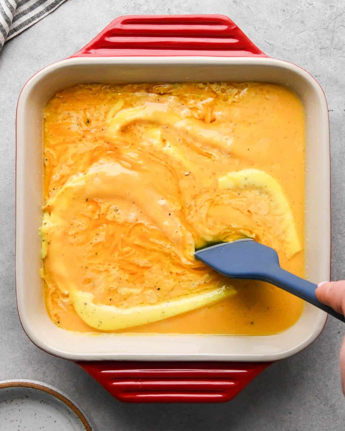 photo showing how to make baked scrambled eggs - a blue spatula stirring the cheese into the partially baked egg mixture in a red, square, ceramic baking dish