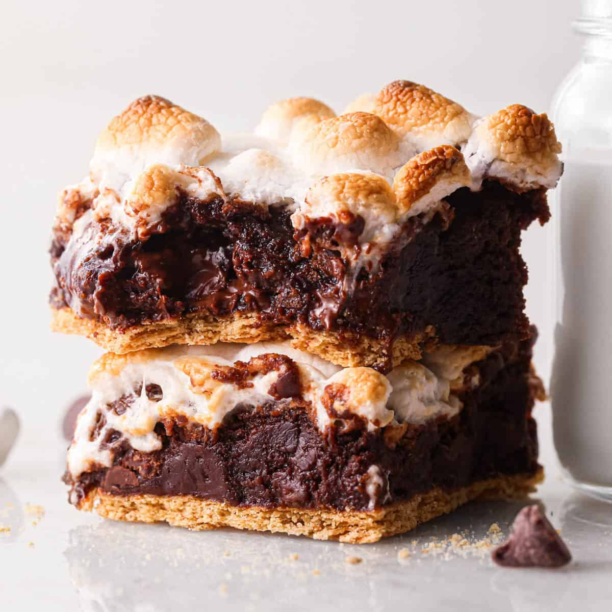 a stack of two s'mores brownies with a graham cracker crust on bottom and toasted marshmallows on top. The top brownie has a bite taken out of it so the fudgy texture of the brownie is visible. There's a glass of milk and chocolate chips in the background