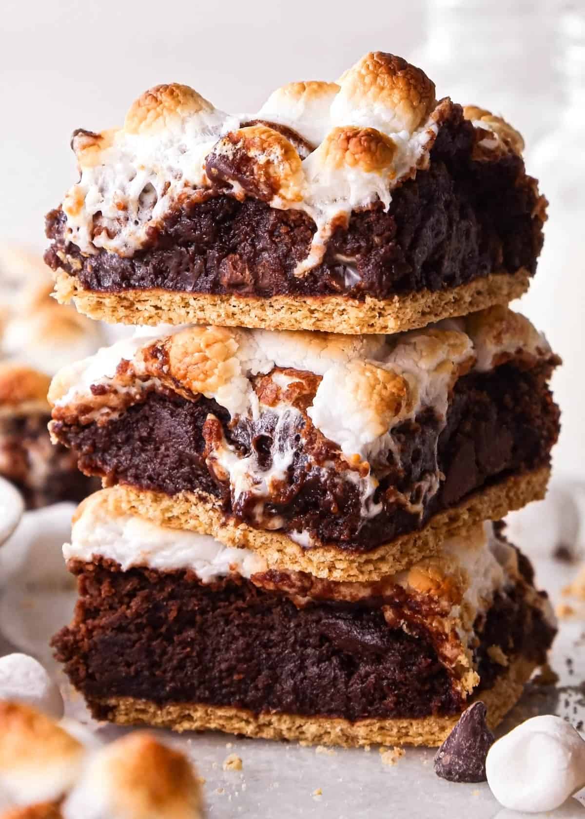 a stack of 3 s'mores brownies with a graham cracker crust on bottom and toasted marshmallows on top. There are toasted marshmallows, chocolate chips and more brownies in the background
