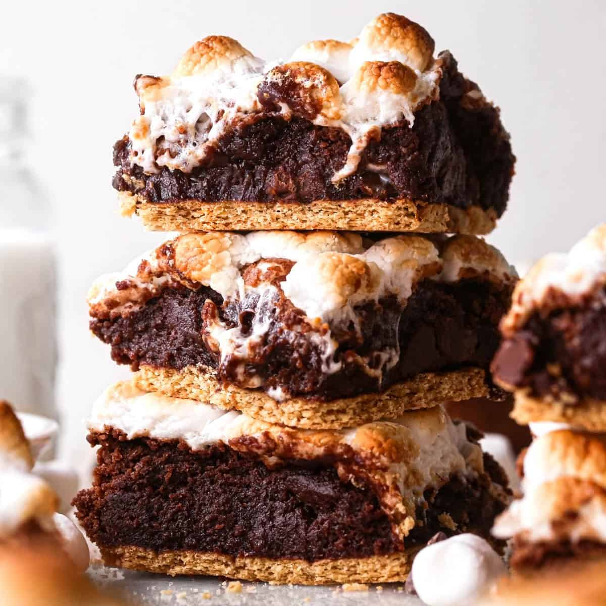 a stack of 3 s'mores brownies with a graham cracker crust on bottom and toasted marshmallows on top. There are toasted marshmallows, chocolate chips and more brownies in the background