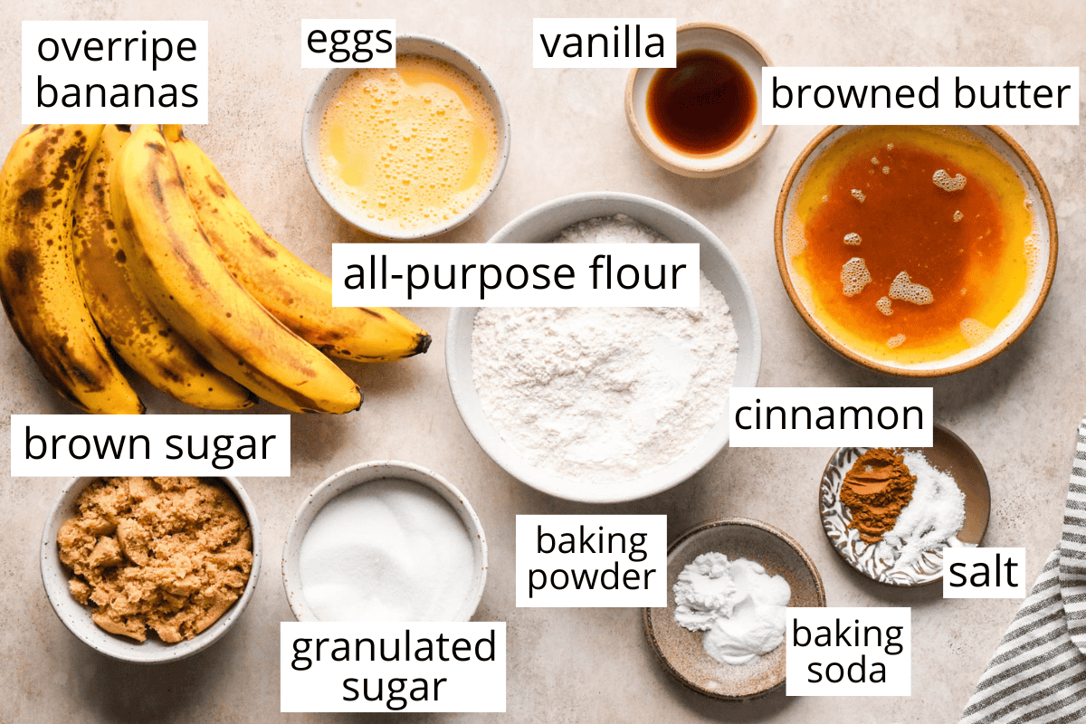 overhead photo of the labeled ingredients in the brown butter banana bread recipe in bowls and labeled like eggs, bananas, browned butter, brown sugar, white sugar, flour, cinnamon salt, vanilla, baking soda and baking powder