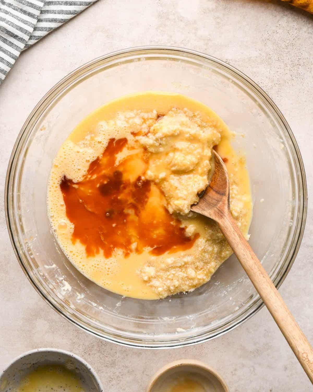 making brown butter banana bread - vanilla and eggs added to the banana mixture in a glass bowl with a wooden spoon