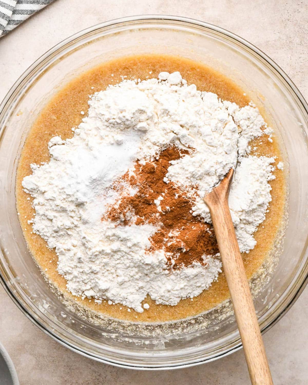 making brown butter banana bread - flour, salt, baking soda, baking powder and cinnamon added to the wet ingredients in a glass bowl with a wooden spoon before mixing