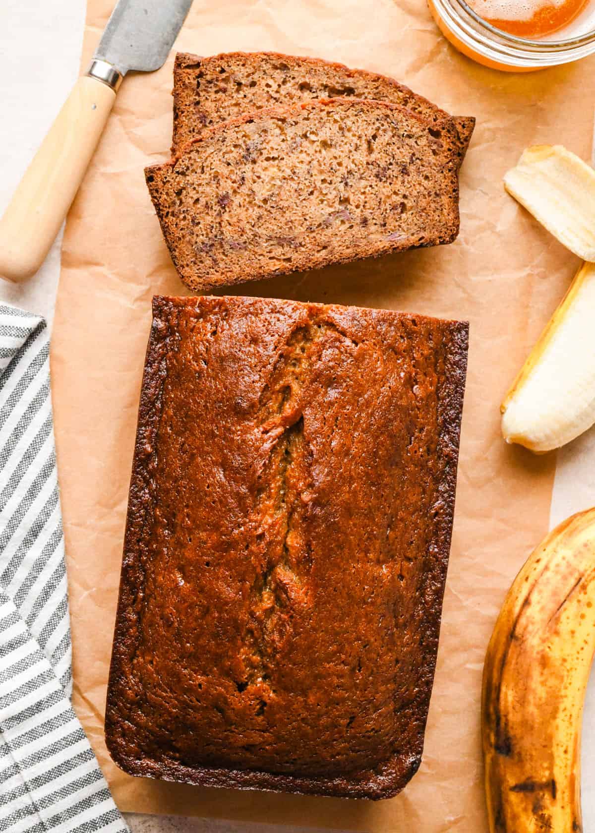 A loaf of Brown butter banana bread on parchment paper with 2 slices cut out of it laying flat showing a moist, tender crumb, surrounded by whole bananas, melted brown butter, and a gray and white striped kitchen towel.