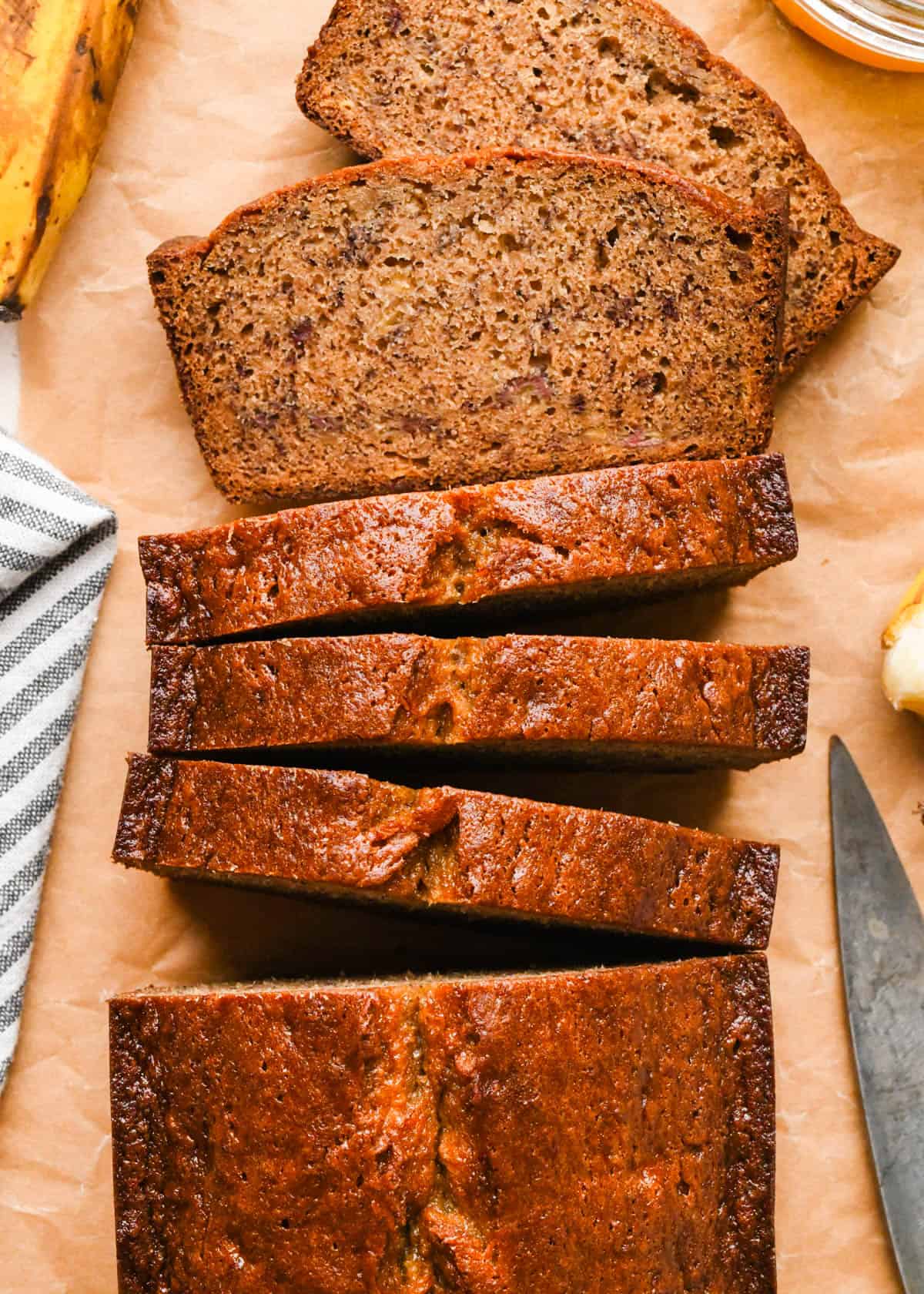 A loaf of Brown butter banana bread with 5 slices cut out of it on parchment paper, two are laying flat showing a moist, tender crumb , surrounded by whole bananas, melted brown butter, and a gray and white striped kitchen towel. and a knife