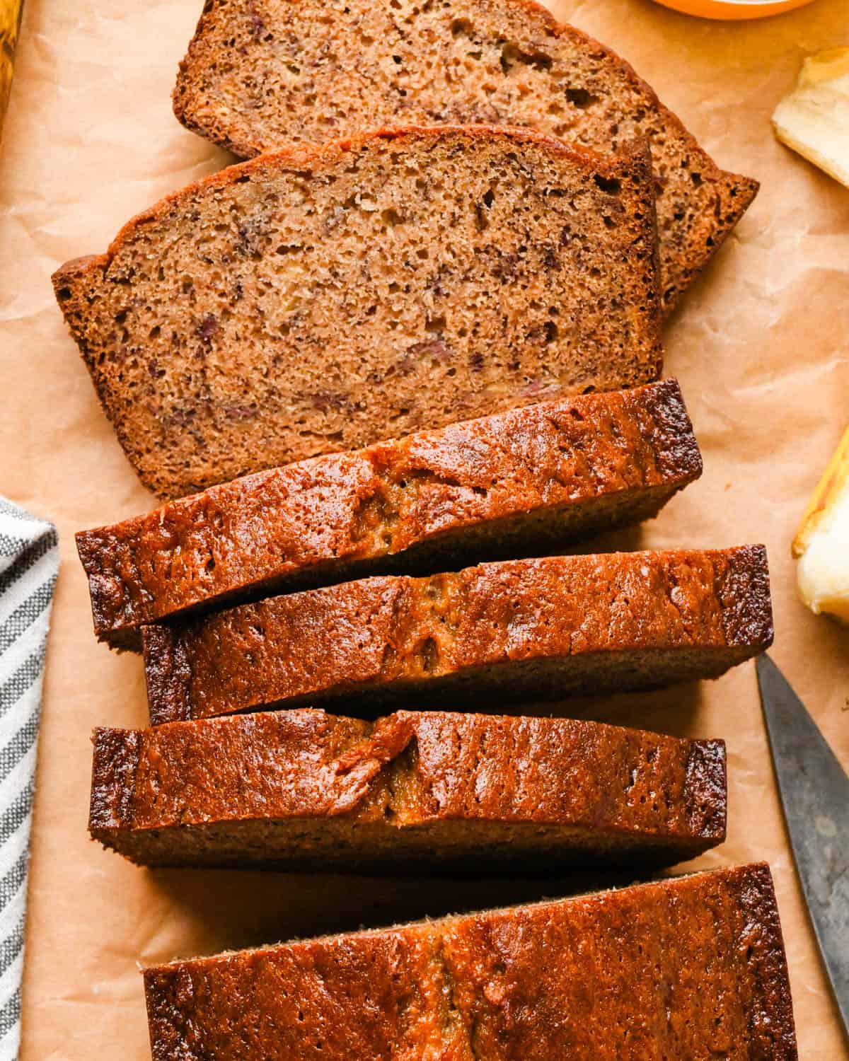 Close up photo of a loaf of Brown butter banana bread with 5 slices cut out of it on parchment paper, two are laying flat showing a moist, tender crumb , surrounded by whole bananas, melted brown butter, and a gray and white striped kitchen towel.