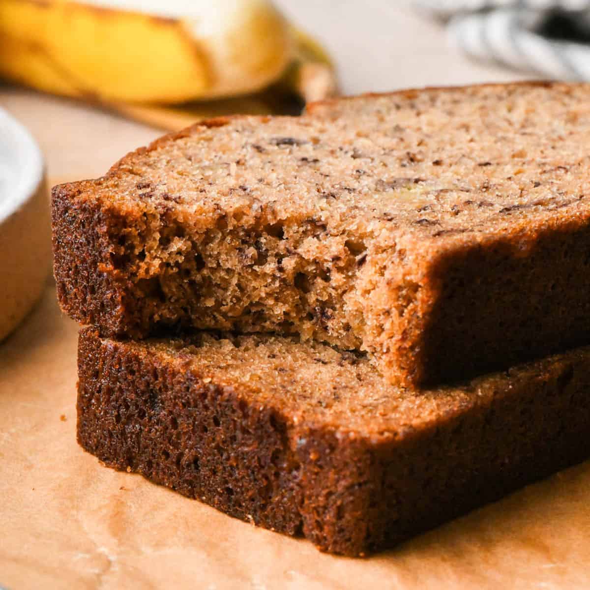 a stack of 2 slices of brown butter banana bread sitting on parchment paper, the top one has a bite taken out of it. There is a banana in the background