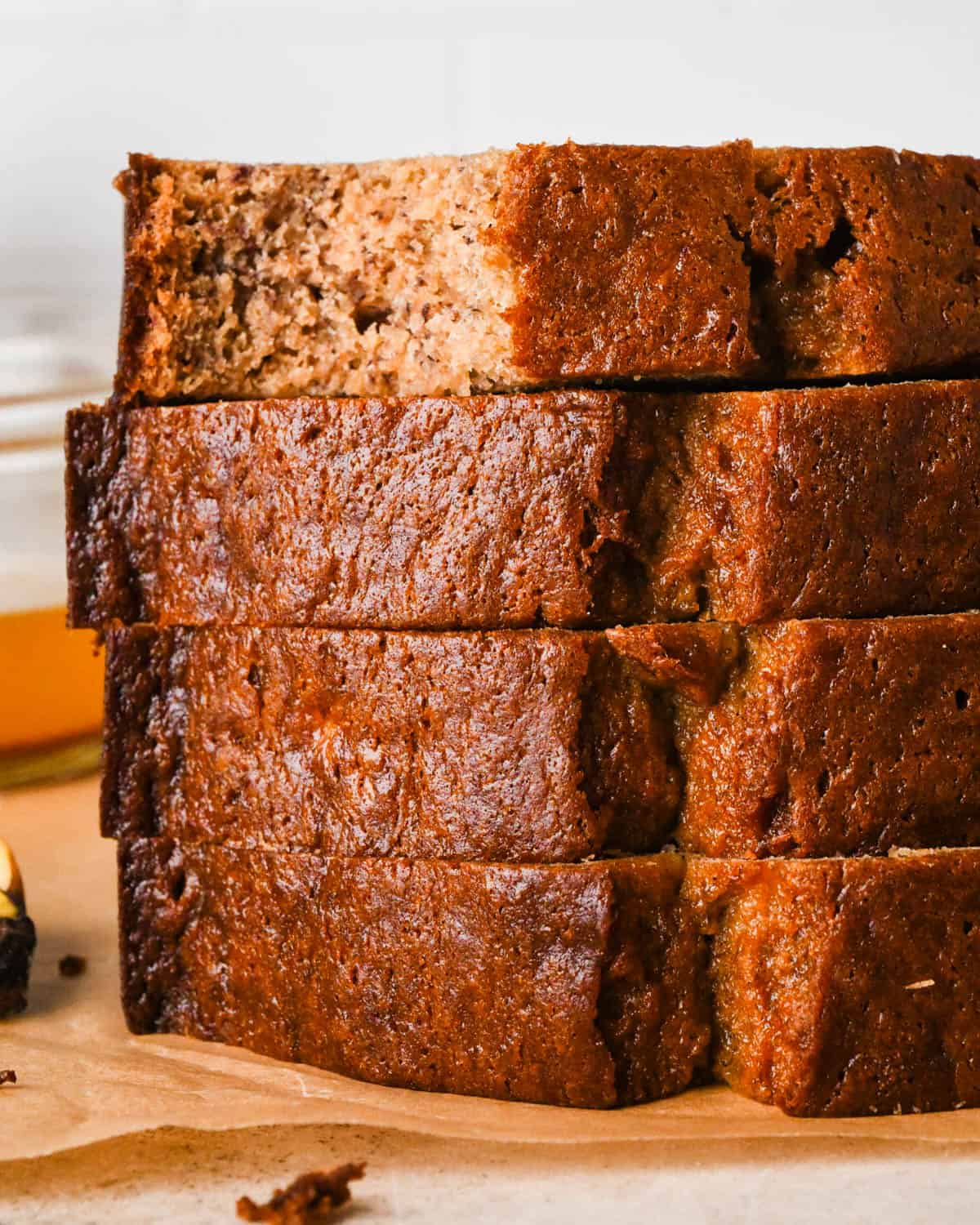 a stack of 4 slices of brown butter banana bread sitting on parchment paper, the top one has a bite taken out of it. There is a banana in the background