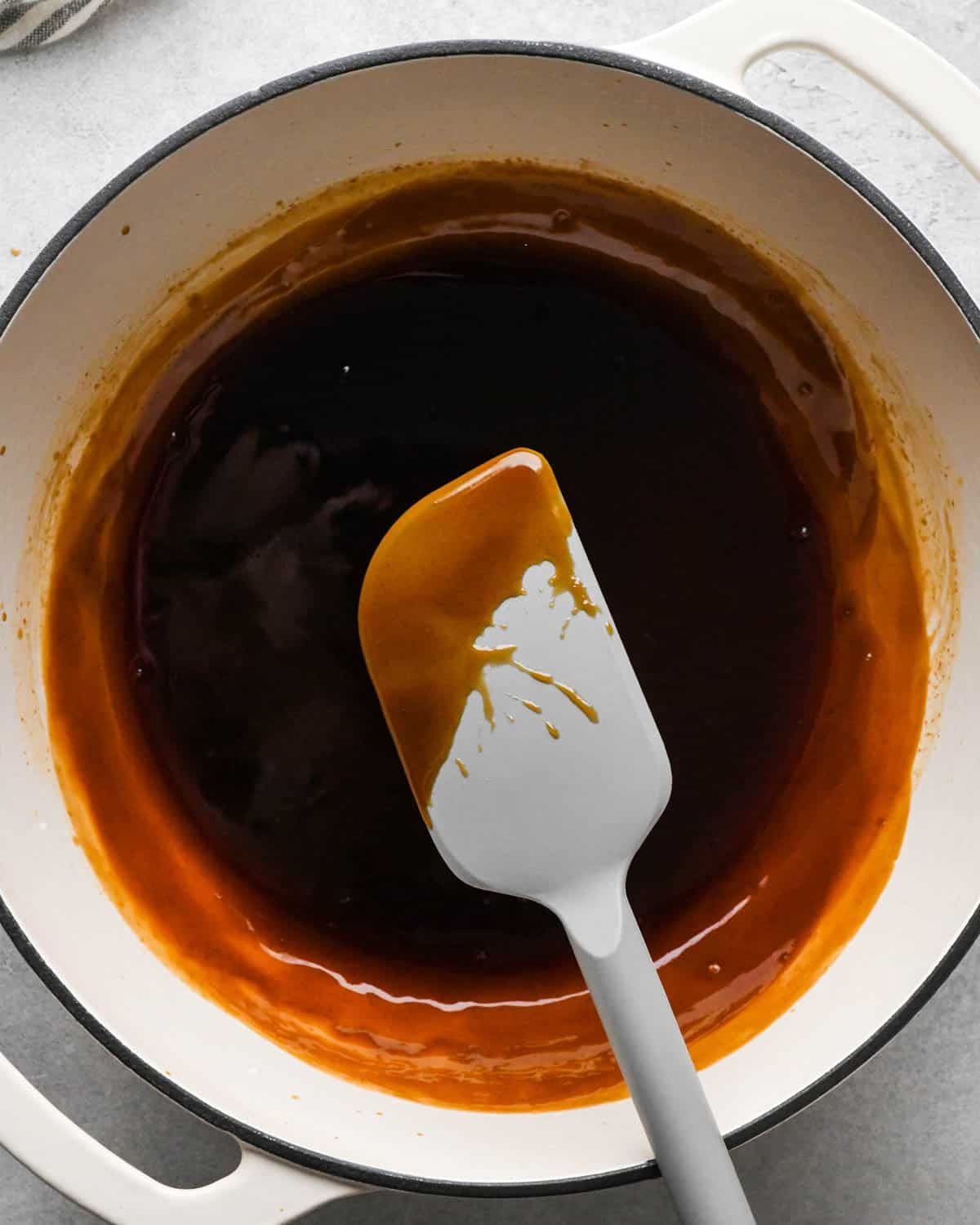 thickened teriyaki sauce in a white saucepan with a black rim, there's a gray spatula with sauce on it showing it has thickened.