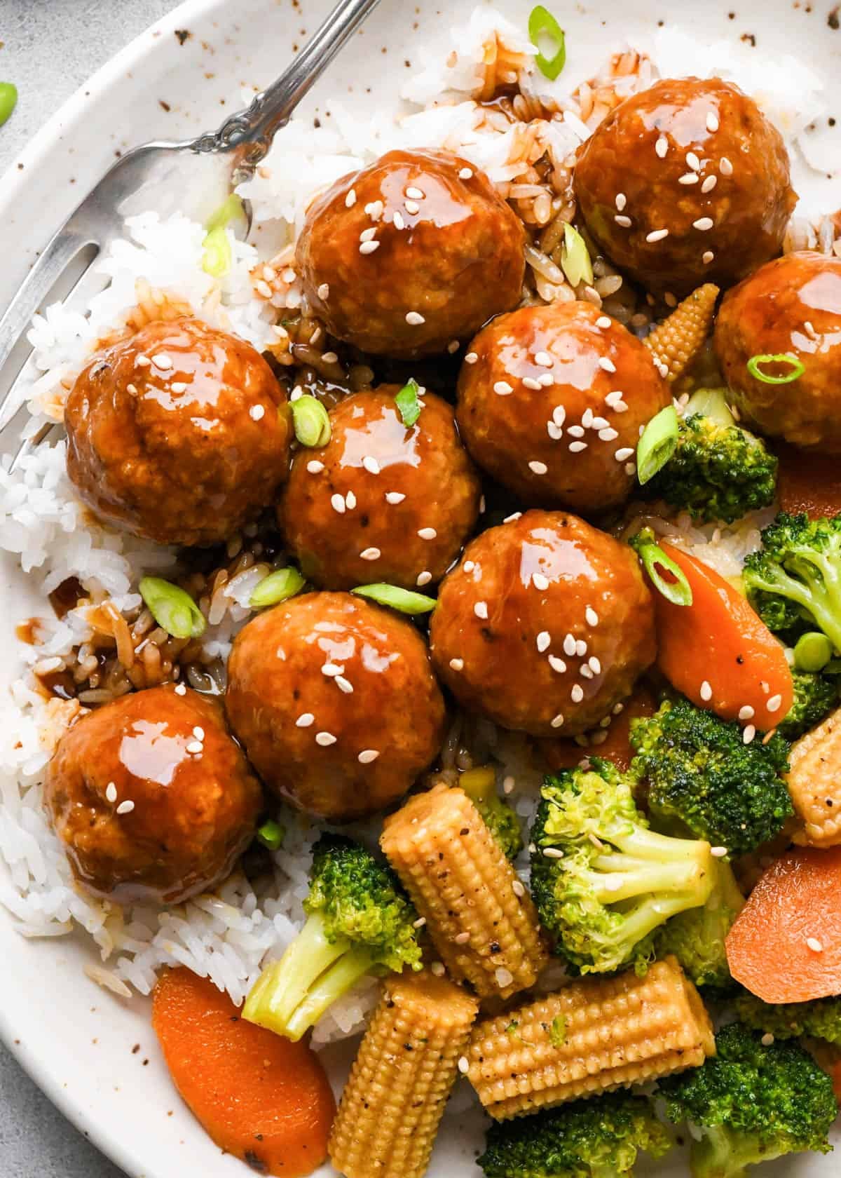 Chicken teriyaki meatballs served over white rice with broccoli, carrots, and baby corn, coated in a homemade teriyaki sauce and topped with sesame seeds and green onions.