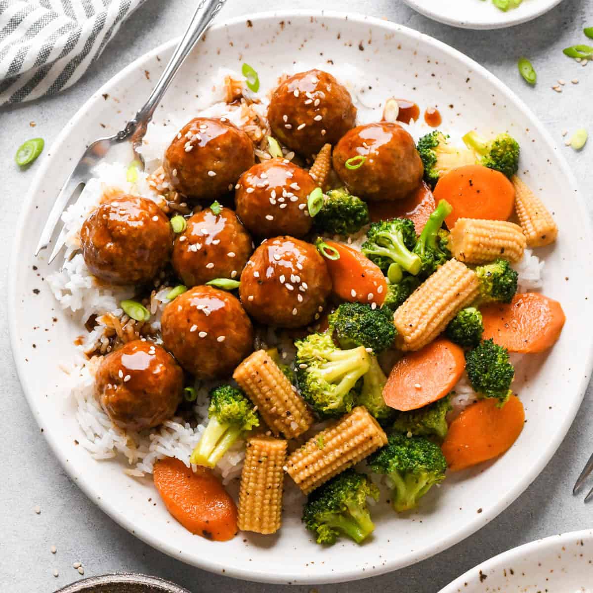 Chicken teriyaki meatballs served over white rice with broccoli, carrots, and baby corn, coated in a homemade teriyaki sauce and topped with sesame seeds and green onions.
