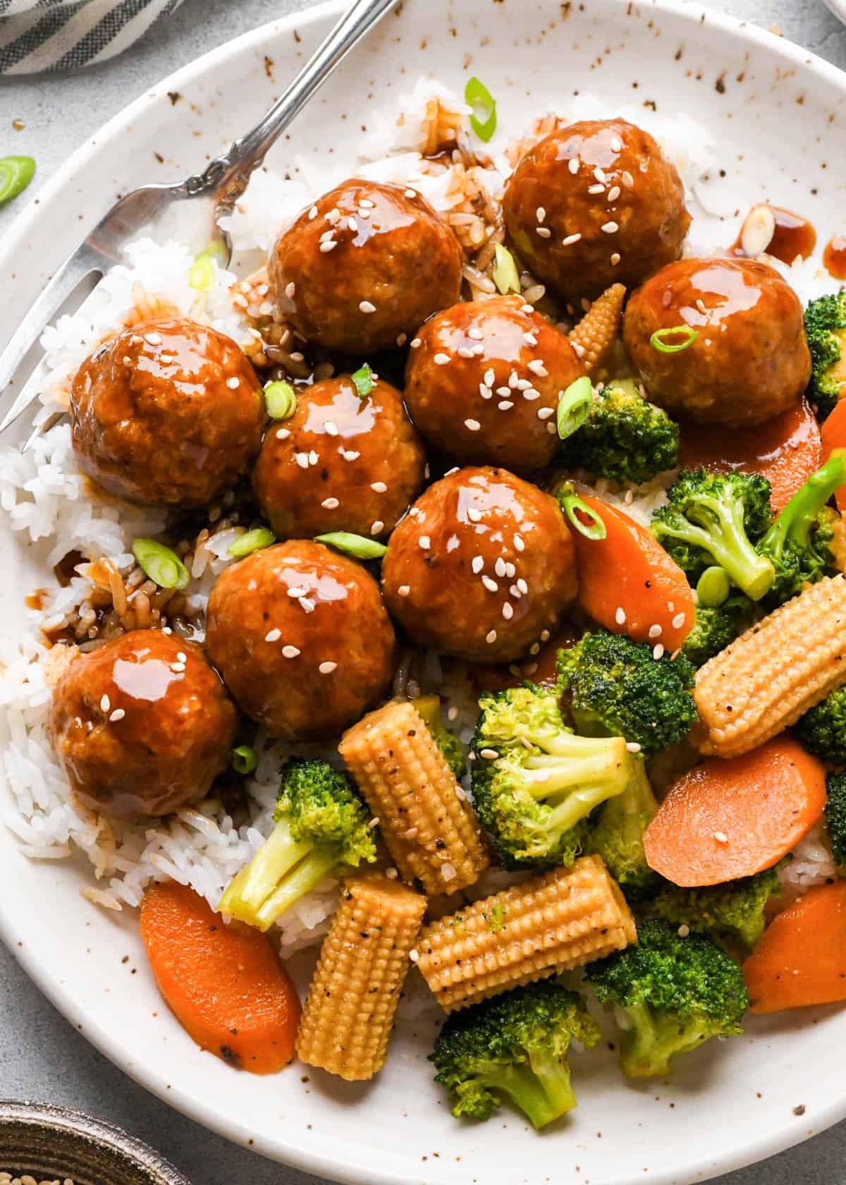 Chicken teriyaki meatballs on a white ceramic plate served over white rice with broccoli, carrots, and baby corn, coated in a homemade teriyaki sauce and topped with sesame seeds and green onions.