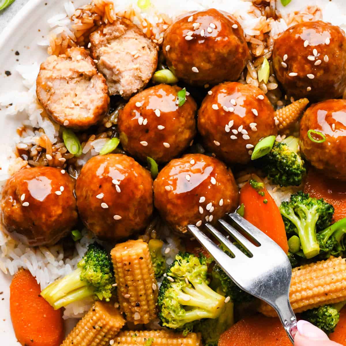 Chicken teriyaki meatballs served over white rice with broccoli, carrots, and baby corn, coated in a homemade teriyaki sauce and topped with sesame seeds and green onions, shown close-up with one meatball cut open to reveal a juicy, tender center. A fork is sticking into one whole meatball