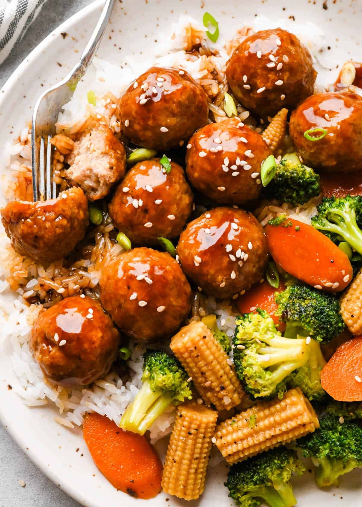 Chicken teriyaki meatballs served over white rice with broccoli, carrots, and baby corn, coated in a homemade teriyaki sauce and topped with sesame seeds and green onions, shown close-up with one meatball cut open to reveal a juicy, tender center