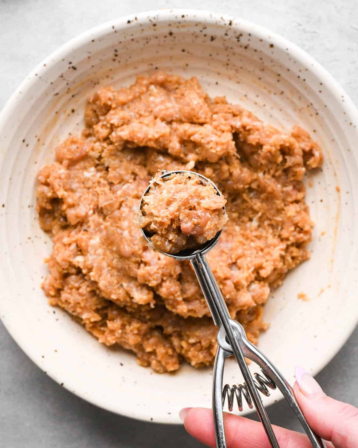 a hand holding a 1 tsp cookie scoop filled with chicken teriyaki meatballs mixture over a white ceramic bowl containing the rest of the mixture.