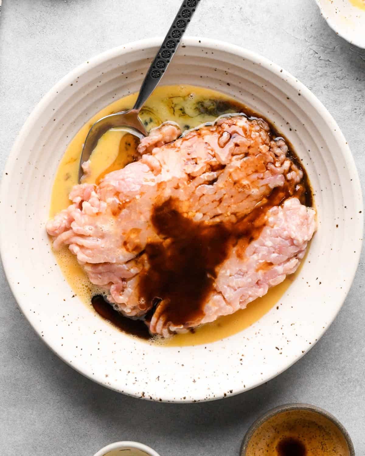 making teriyaki meatballs - ground chicken, egg, soy sauce, hoisin sauce, and sesame oil in a ceramic mixing bowl with a metal spoon before mixing