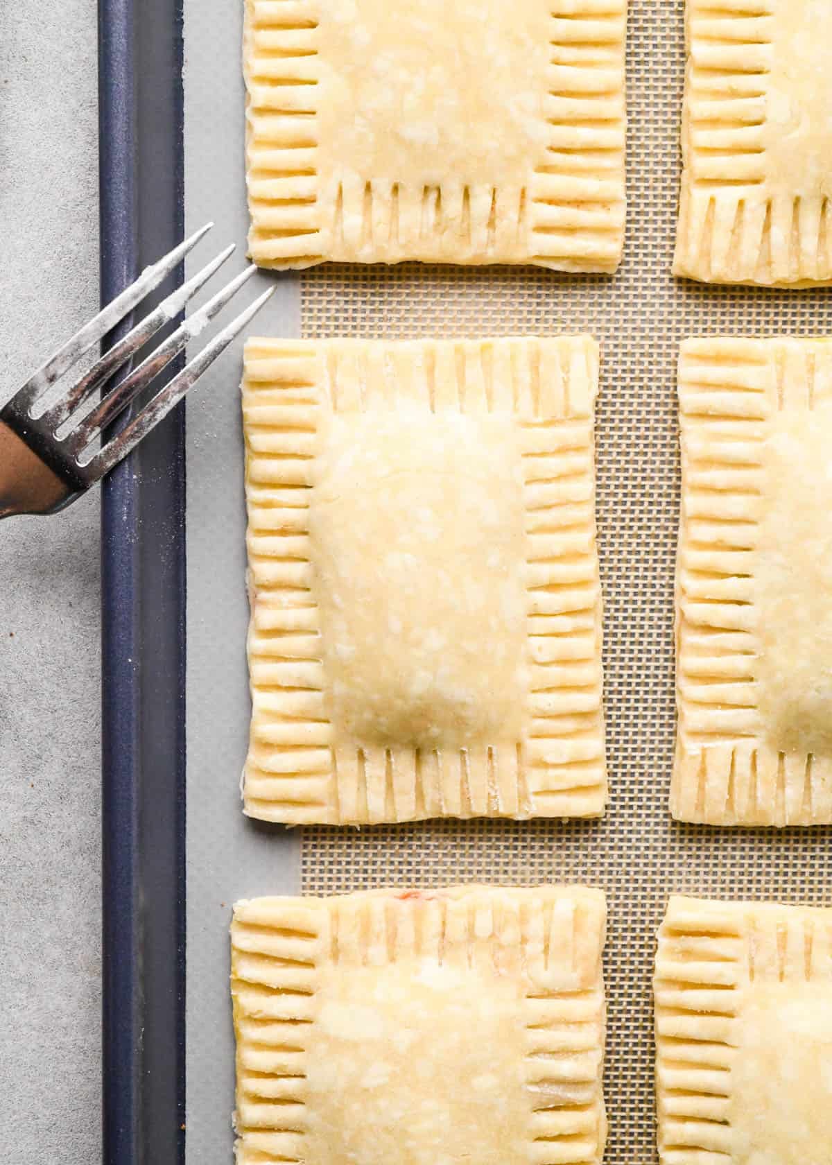 making homemade pop tarts - a fork crimping the edges of 6 pop tarts on a baking sheet lined with a silicone baking mat before baking