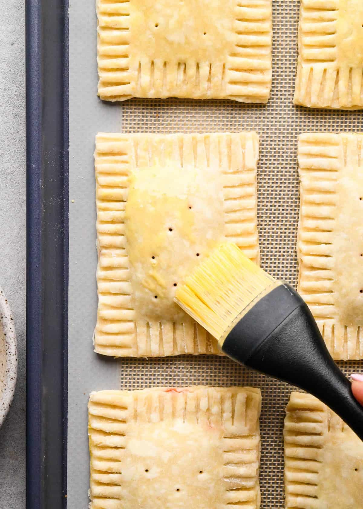 making homemade pop tarts - egg wash being brushed on 6 pop tarts with holes punched into the top on a baking sheet lined with a silicone baking mat before baking