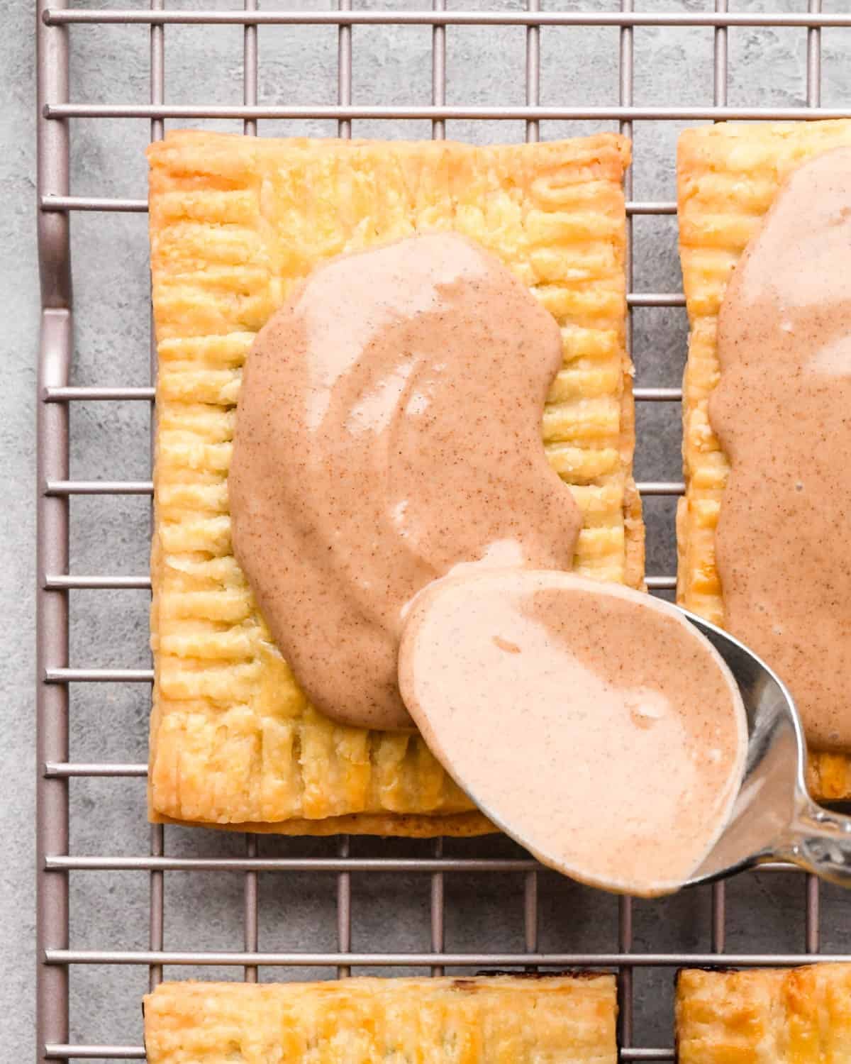 making homemade pop tarts - cinnamon icing being spread on top of a pop tart with a metal spoon. The pop tart is on a wire cooling rack with 3 other pop tarts around it