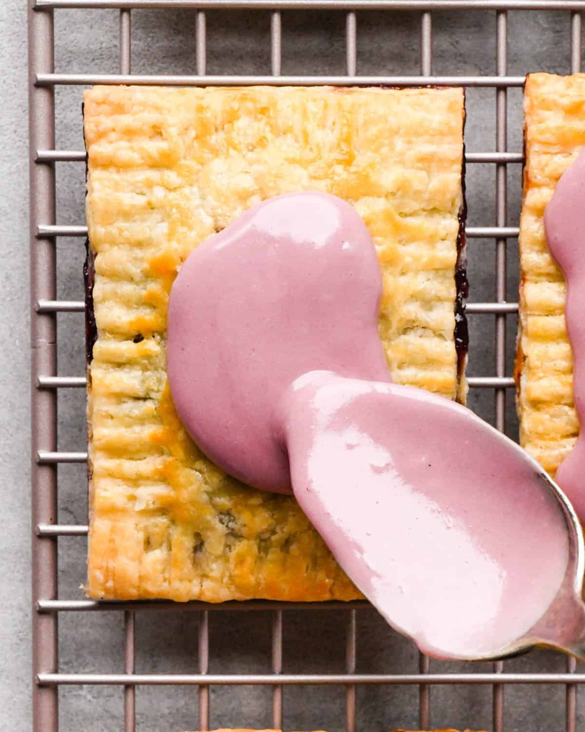 making homemade pop tarts - blackberry icing being spread on top of a pop tart with a metal spoon. The pop tart is on a wire cooling rack with 3 other pop tarts around it