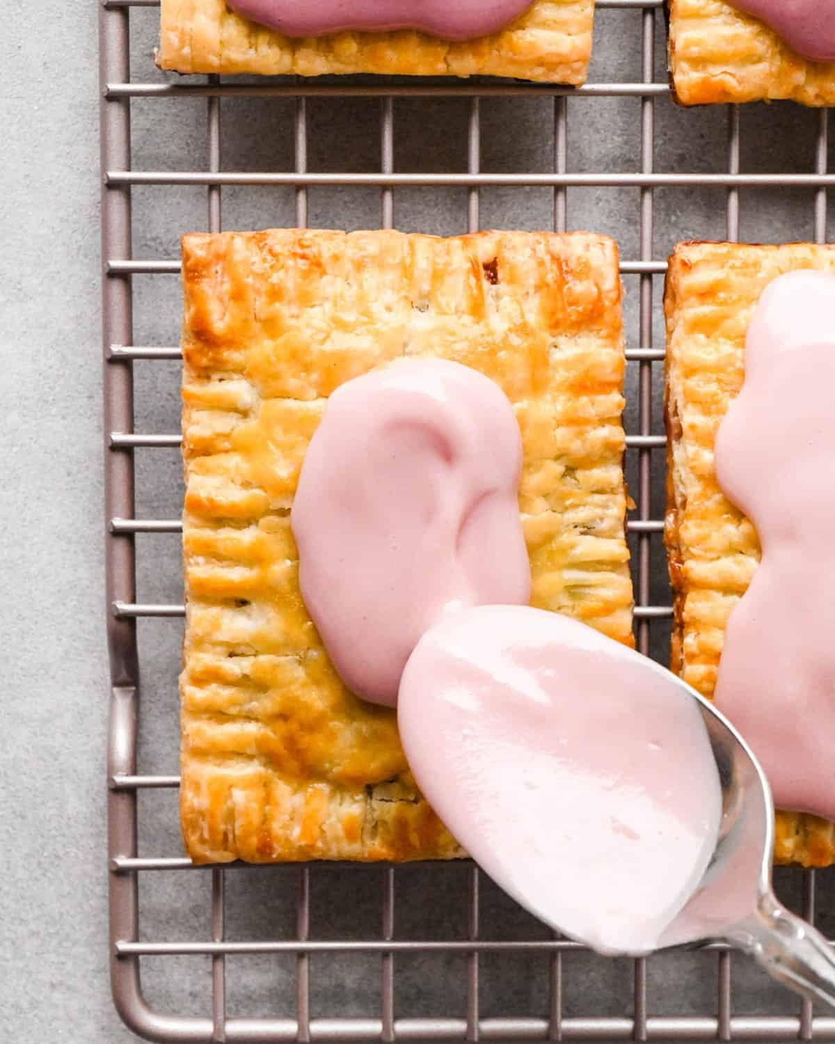 making homemade pop tarts - strawberry icing being spread on top of a pop tart with a metal spoon. The pop tart is on a wire cooling rack with 3 other pop tarts around it