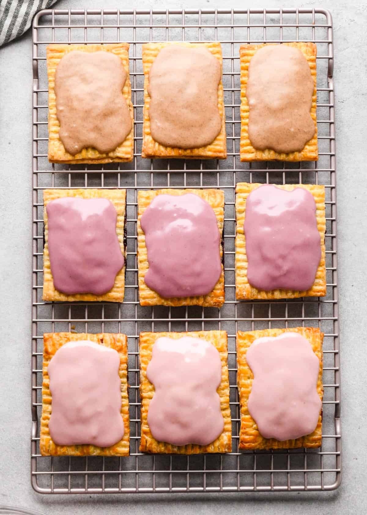 9 homemade pop tarts in 3 rows of 3 on a metal cooling rack. The top row is 3 cinnamon sugar pop tarts, the middle row is 3 blackberry pop tarts and the bottom row is three strawberry pop tarts. All with icing on top.