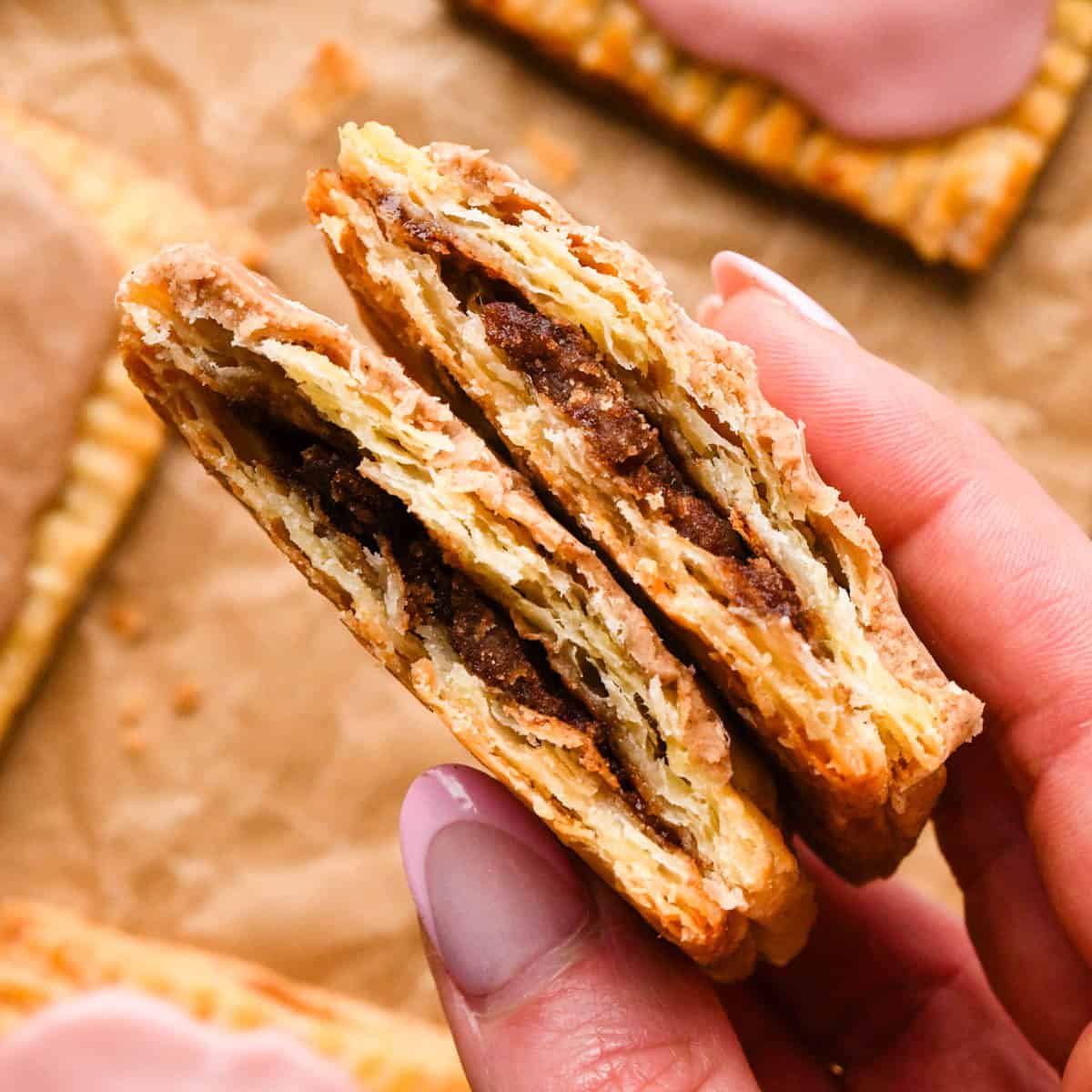 up close photo of a two halves of a cinnamon sugar homemade pop tart resting on top of each other so the cinnamon filling is visible with a cinnamon glaze on top, with 3 other homemade pop tarts in the background all on parchment paper