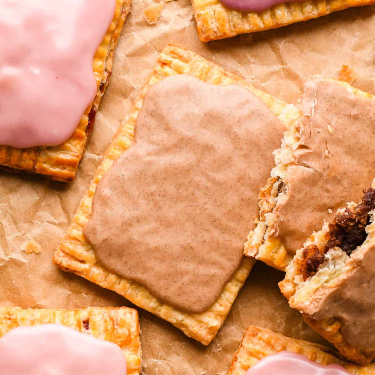 5 Homemade pop tarts, 2 cinnamon sugar and 3 strawberry, topped with pink strawberry and cinnamon glaze, one broken open to reveal cinnamon filling on parchment paper.