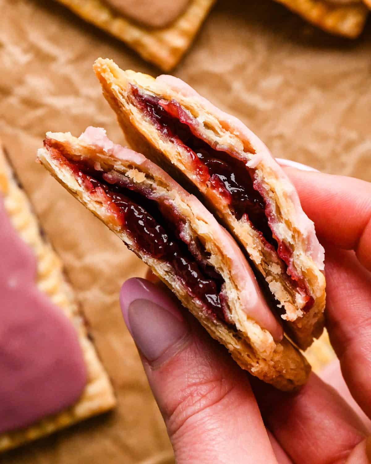 a hand holding two halves of a strawberry homemade pop tart resting on top of each other so the strawberry filling is visible with a pink strawberry glaze on top
