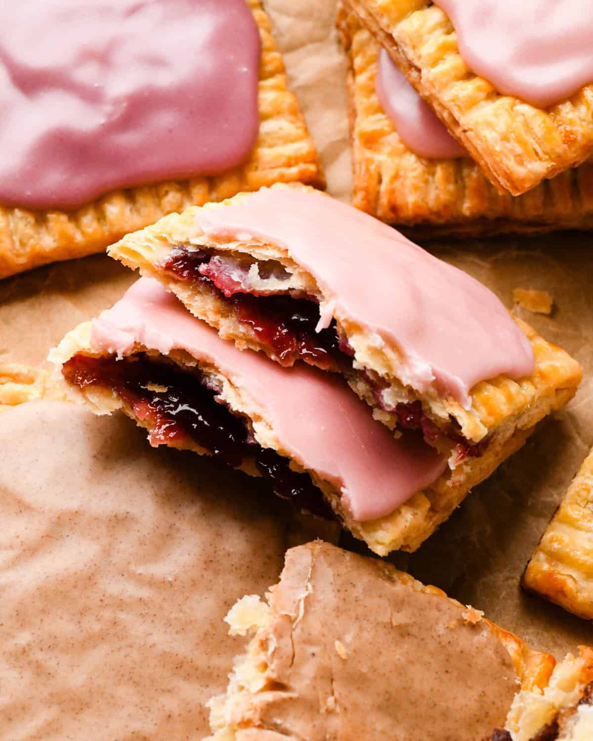 up close photo of a two halves of a strawberry homemade pop tart resting on top of each other so the strawberry filling is visible with a pink strawberry glaze on top, with 4 other homemade pop tarts around them all on parchment paper