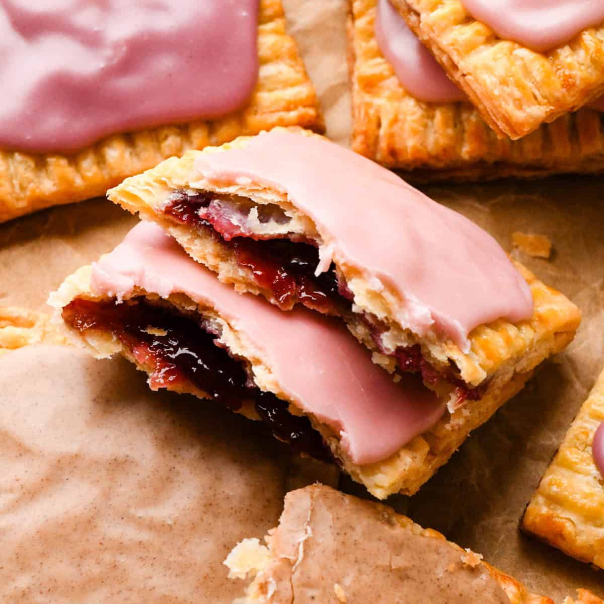 two halves of a strawberry homemade pop tart resting on top of each other so the strawberry filling is visible with a pink strawberry glaze on top, with 4 other homemade pop tarts around them all on parchment paper