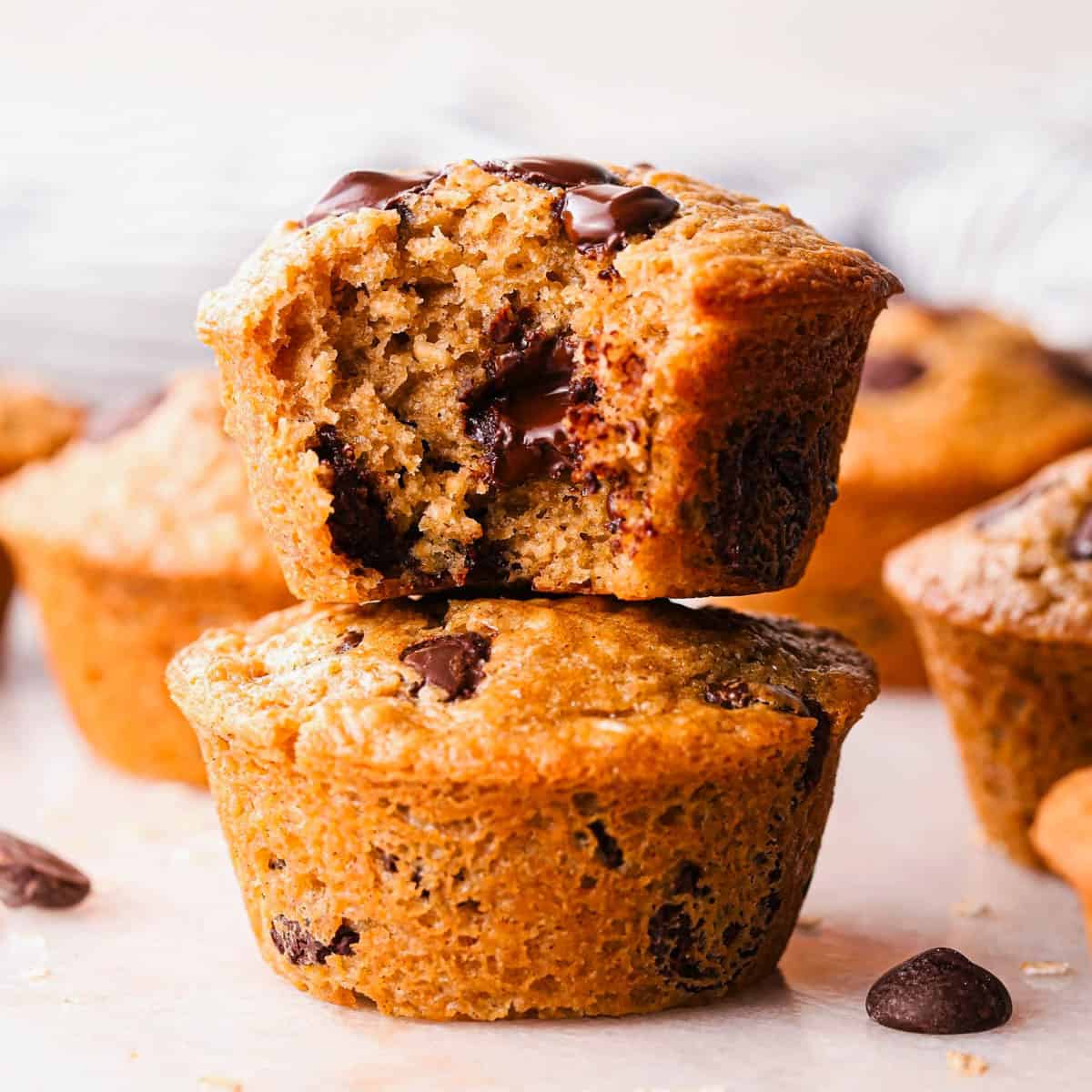 a stack of 2 oatmeal muffins chocolate chip muffins, the top one has a bite taken out of it so the fluffy texture and melty chocolate chips inside are visible. Surrounded by more muffins, and more chocolate chips