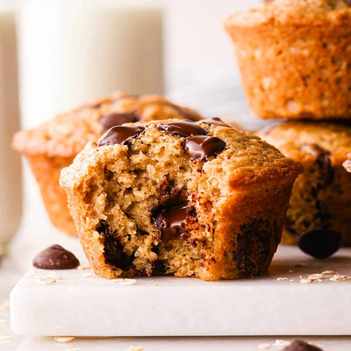 4 oatmeal muffins chocolate chip muffins, one has a bite taken out of it so the fluffy texture and melty chocolate chips inside are visible. a bottle of milk and more chocolate chips in the background