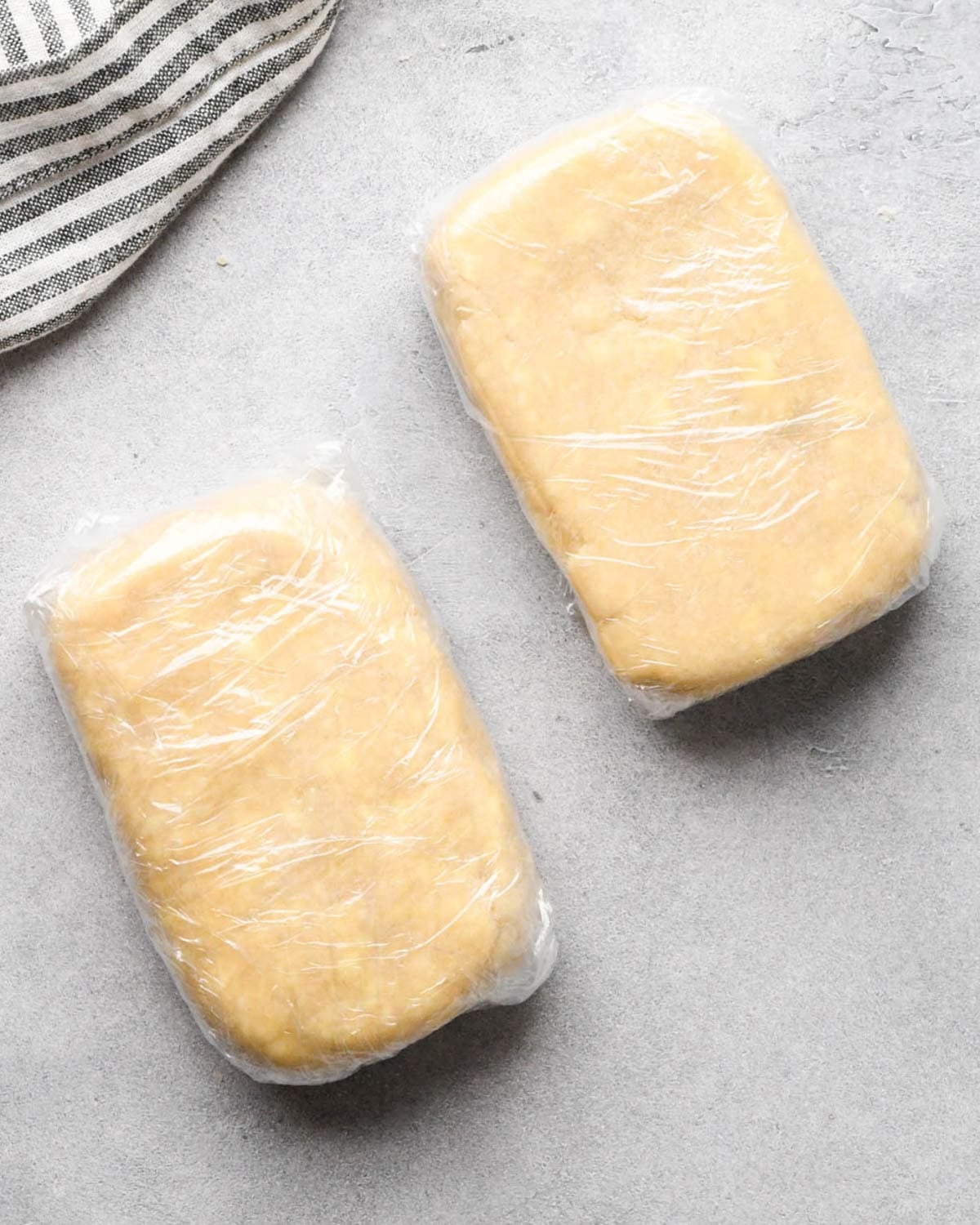 making apple hand pies crust - two rectangles of dough wrapped in plastic wrap on a gray surface, there is a striped towel in the top left corner