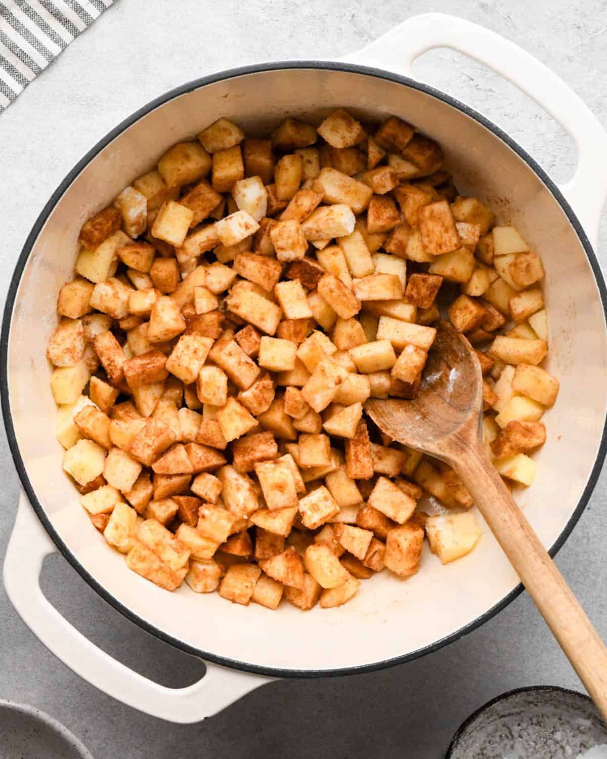 making apple hand pies filling - diced and peeled apples, cinnamon, brown sugar, granulated sugar, cornstarch, vanilla and nutmeg in a white pot during cooking with a wooden spoon