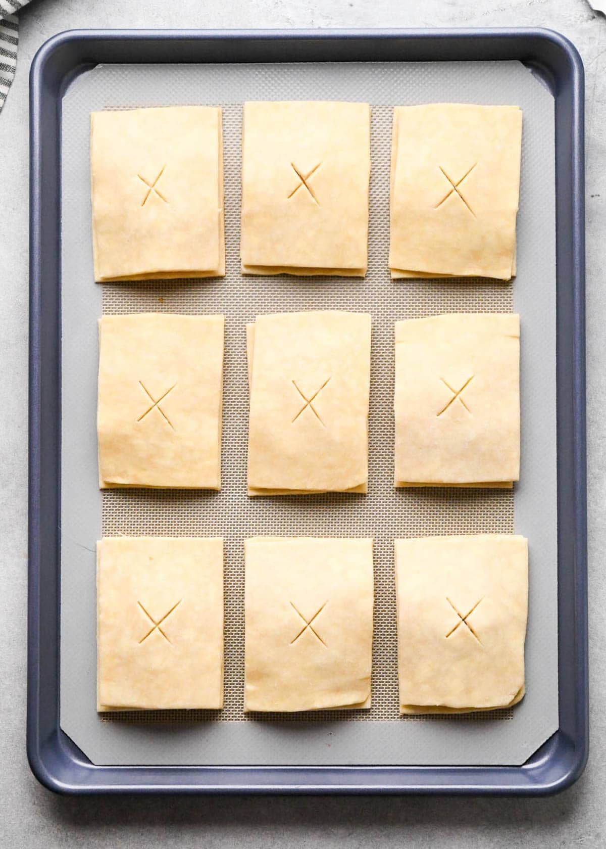 9 assembled apple hand pies on a parchment lined baking sheet with an "X" cut into the center of each one