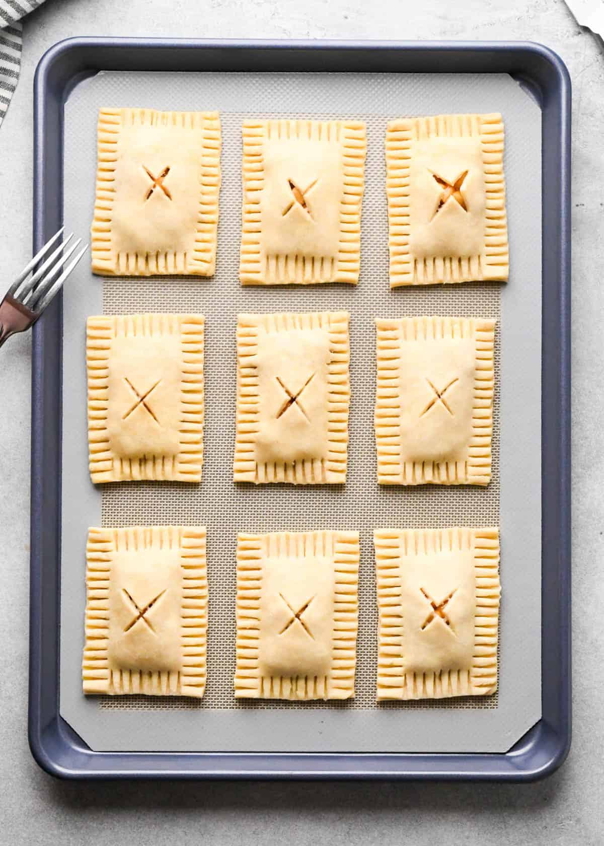 9 assembled apple hand pies on a parchment lined baking sheet with an "X" cut into the center of each one and now they all have crimped edges and there's a fork on the left hand side.
